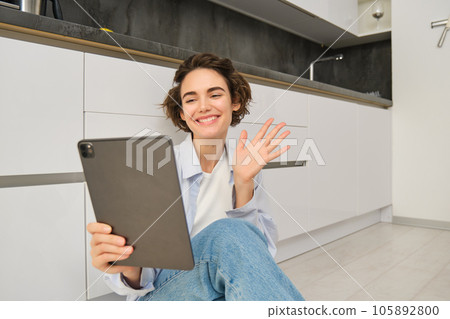 Portrait of happy young woman connects to video chat, waves at digital tablet and smiling, sits at home Portrait of happy young woman connects to video chat, waves at digital tablet and smiling, sits at home 105892800