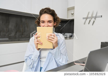 Portrait of smiling brunette girl, holds her planner, writes in notebook and looks happy, laughs, does homework, makes notes, sits at home in kitchen 105892828
