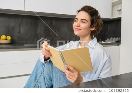 Portrait of young adult woman, reading her notes, holding notebook and smiling, relaxing at home Portrait of young adult woman, reading her notes, holding notebook and smiling, relaxing at home 105892835
