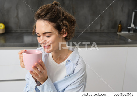 Portrait of a joyful young woman enjoying a cup of coffee at home. Smiling beautiful girl drinks hot tea in morning. Excited woman laughing on an autumn day 105892845