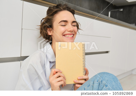 Portrait of happy, carefree woman, writes in her journal and laughs, holds her diary, reads something in notebook, sits on floor indoors 105892869