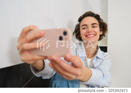 Portrait of beautiful brunette girl, takes selfie on smartphone in her kitchen, posing for photo on mobile phone Portrait of beautiful brunette girl, takes selfie on smartphone in her kitchen, posing for photo on mobile phone 105892871