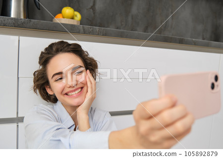 Portrait of happy, beautiful woman takes selfie on smartphone, extends her hand with mobile phone and takes picture of herself, records vlog on mobile phone 105892879
