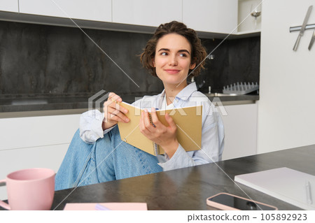 Young woman sitting in kitchen, reading her notes in notebook. Smiling girl at home, doing homework, studying Young woman sitting in kitchen, reading her notes in notebook. Smiling girl at home, doing homework, studying 105892923