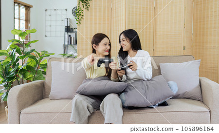 Asian beautiful lesbian gay couple enjoy play game together in Livingroom. Two female friend sit on sofa in living room, feel happy to play joystick game at home. 105896164