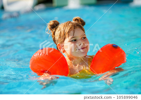 Little preschool girl with protective swimmies playing in outdoor swimming pool by sunset. Child learning to swim in outdoor pool, splashing with water, laughing and having fun. Family vacations. Little preschool girl with protective swimmies playing in outdoor swimming pool by sunset. Child learning to swim in outdoor pool, splashing with water, laughing and having fun. Family vacations. 105899046