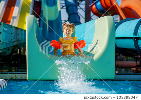 Little preschool girl on water slide in aqua park. Happy child having fun on water slides on family summer vacation in tropical resort. Amusement park with wet playground for young child and baby. Little preschool girl on water slide in aqua park. Happy child having fun on water slides on family summer vacation in tropical resort. Amusement park with wet playground for young child and baby. 105899047