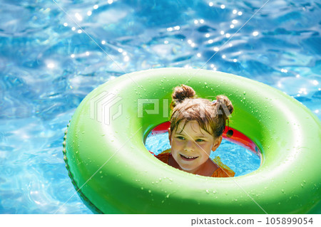 Happy little girl with inflatable toy ring float in swimming pool. Little preschool child learning to swim and dive in outdoor pool of hotel resort. Healthy sport activity and fun for children. Happy little girl with inflatable toy ring float in swimming pool. Little preschool child learning to swim and dive in outdoor pool of hotel resort. Healthy sport activity and fun for children. 105899054