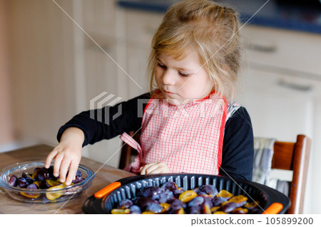 Cute little toddler girl baking plum pie at home. Happy smiling child helping and preparing plums for cake in domestic kitchen. Healthy homemade food. 105899200