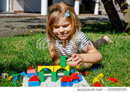 Little preschool girl playing with wooden blocks on the table outdoors. Educational game for small children. Happy child builds a tower from wooden rainbow stacking blocks. 105899595