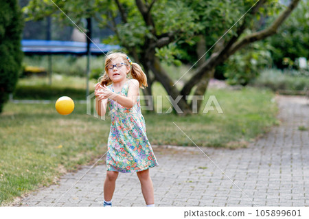 Little preschool girl with eyeglasses playing with ball outdoors. Happy smiling child catching and throwing, laughing and making sports. Active leisure with children and kids. Summer day on backyard Little preschool girl with eyeglasses playing with ball outdoors. Happy smiling child catching and throwing, laughing and making sports. Active leisure with children and kids. Summer day on backyard 105899601