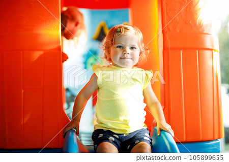 Happy blond little toddler girl having fun and sliding on indoor playground at daycare or nursery. Positive funny baby child smiling. Healthy girl climbing on slide. 105899655