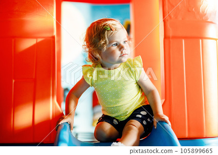 Happy blond little toddler girl having fun and sliding on indoor playground at daycare or nursery. Positive funny baby child smiling. Healthy girl climbing on slide. 105899665