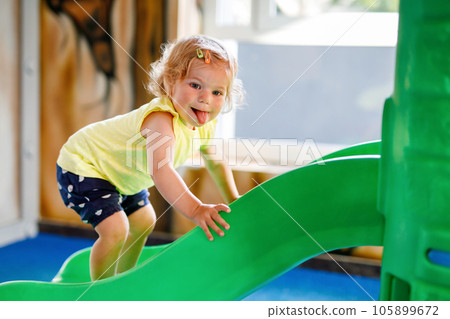 Happy blond little toddler girl having fun and sliding on indoor playground at daycare or nursery. Positive funny baby child smiling. Healthy girl climbing on slide. 105899672