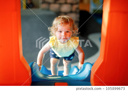 Happy blond little toddler girl having fun and sliding on indoor playground at daycare or nursery. Positive funny baby child smiling. Healthy girl climbing on slide. 105899673