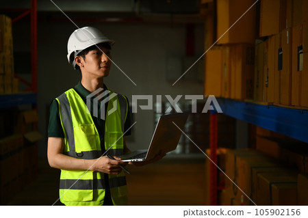 Male manager in hardhat and vest using laptop, checking inventory production stock control in warehouse 105902156