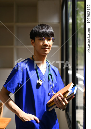 Smiling asian male medical student man wearing blue scrubs standing near window and looking at camera Smiling asian male medical student man wearing blue scrubs standing near window and looking at camera 105902162