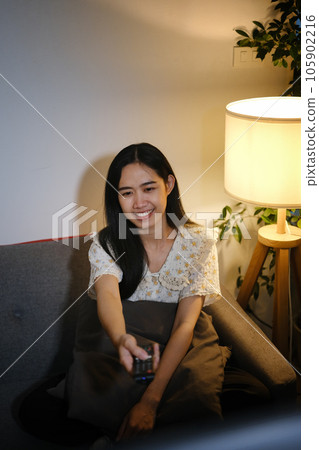 Beautiful young Asian woman having free time watching tv, holding remote control switch channels 105902216