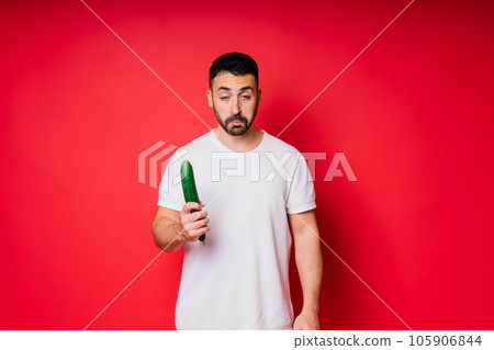 Young man in white shirt isolated over background. Guy look at long cucumber in hand. 105906844