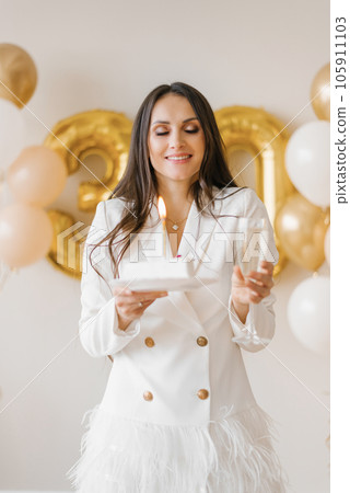 Young caucasian woman holding a cake with a candle and a glass of champagne in honor of the thirtieth birthday in a stylish white dress with feathers 105911103