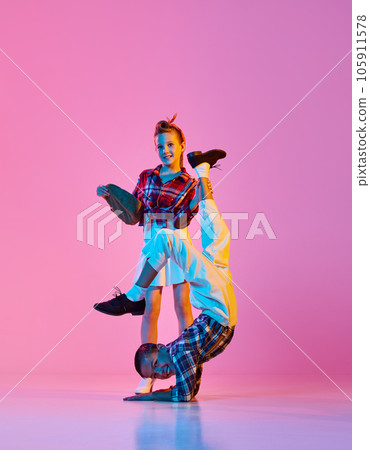 Flexibility. Stylish, active children, girl and boy in retro style clothes dancing against pink studio background in neon light 105911578