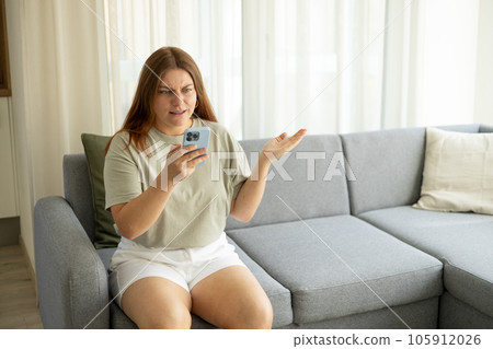 Upset young woman using phone and reading bad news, sad pensive girl sitting at home. Negative people emotion. Redhead woman holding smartphone, using cellphone modern technology, looking at mobile Upset young woman using phone and reading bad news, sad pensive girl sitting at home. Negative people emotion. Redhead woman holding smartphone, using cellphone modern technology, looking at mobile 105912026