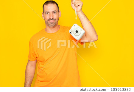 Hispanic man with a beard in his 40s with a photo camera hanging, isolated on a yellow studio background. 105913883