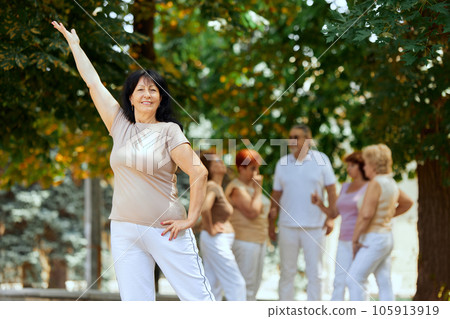 Focus on senior cheerful woman standing in park on warm sunny day. Blurred people on background 105913919