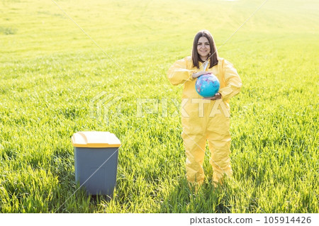 A woman in a yellow protective suit stands in the middle of a green field and holds a globe in her hands, next to a garbage can 105914426