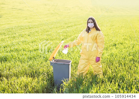 A woman in a yellow protective suit stands in the middle of a green field and holds a plastic bottle in her hands to throw in the tank A woman in a yellow protective suit stands in the middle of a green field and holds a plastic bottle in her hands to throw in the tank 105914434
