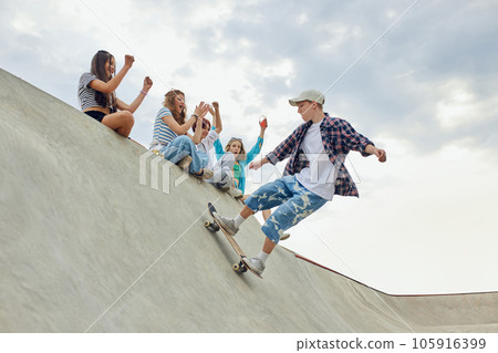 Friends, teengaers meeting at skate park for active leisure time. Boy in casucl clothes skateboarding on ramp 105916399