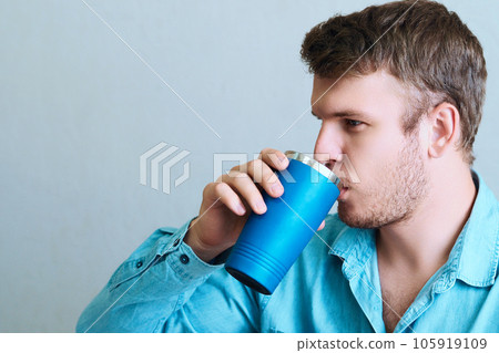Young man drinks drink or medicine from thermo mug. Portrait of man from side on gray background 105919109