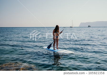 Woman sea sup. Close up portrait of happy young caucasian woman with long hair looking at camera and smiling. Cute woman portrait in a blue bikini posing on sup board in the sea Woman sea sup. Close up portrait of happy young caucasian woman with long hair looking at camera and smiling. Cute woman portrait in a blue bikini posing on sup board in the sea 105921358