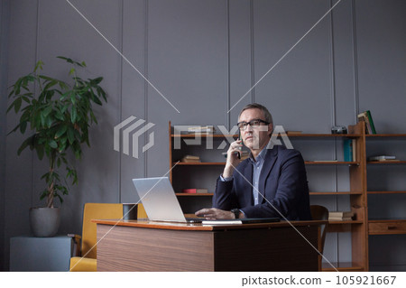Elderly businessman in glasses working on laptop and talking on mobile phone Elderly businessman in glasses working on laptop and talking on mobile phone 105921667