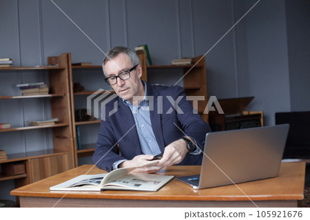 Portrait of adult businessman in glasses and blue suit sitting by laptop and reading the book 105921676