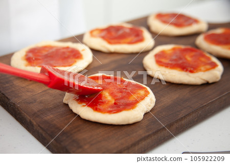 Senior woman adding pizza sauce to the stretched dough. Delicious homemade mini pizzas preparation. Senior woman adding pizza sauce to the stretched dough. Delicious homemade mini pizzas preparation. 105922209