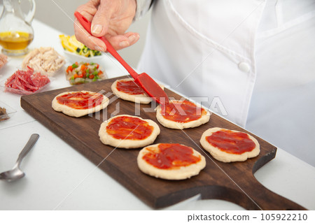 Senior woman adding pizza sauce to the stretched dough. Delicious homemade mini pizzas preparation. Senior woman adding pizza sauce to the stretched dough. Delicious homemade mini pizzas preparation. 105922210