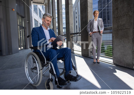 Serious business man in wheelchair with tablet, woman partner standing nearby. Serious business man in wheelchair with tablet, woman partner standing nearby. 105922519