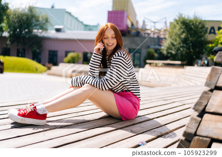 Portrait of happy relaxed young woman wearing skirt sitting resting on wooden bench in public park on summer sunny day, talking using smartphone. Smiling redhead female making call on mobile phone Portrait of happy relaxed young woman wearing skirt sitting resting on wooden bench in public park on summer sunny day, talking using smartphone. Smiling redhead female making call on mobile phone 105923299