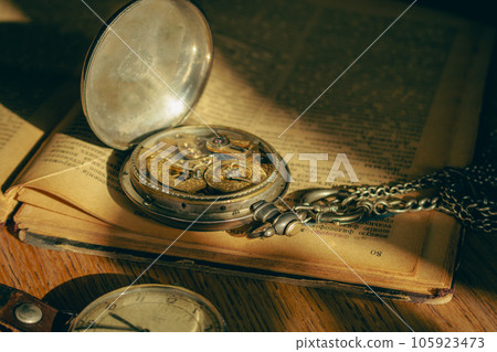 Antique pocket watch and ancient open book. Retro style objects. Vintage silver clock with chain on book. Nostalgia concept. The past background. Watch mechanism close up. Literature background. Antique pocket watch and ancient open book. Retro style objects. Vintage silver clock with chain on book. Nostalgia concept. The past background. Watch mechanism close up. Literature background. 105923473