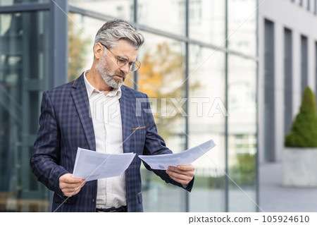 Serious thinking businessman boss with documents contracts invoices outside office building, mature experienced man in business suit looking at papers, with graphs financial reports. Serious thinking businessman boss with documents contracts invoices outside office building, mature experienced man in business suit looking at papers, with graphs financial reports. 105924610