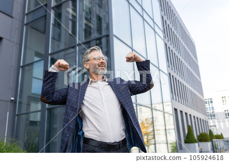 Successful and happy gray-haired boss outside office building rejoices success and victory, triumphs with hands up, businessman business owner in business suit and glasses, mature investor banker. Successful and happy gray-haired boss outside office building rejoices success and victory, triumphs with hands up, businessman business owner in business suit and glasses, mature investor banker. 105924618