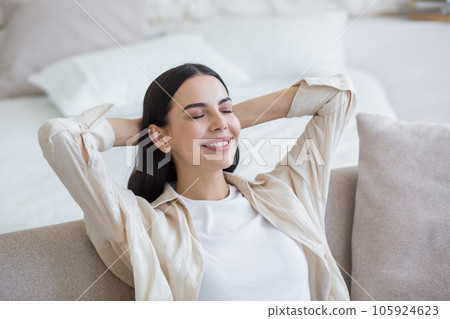 Beautiful young smiling woman relaxing on sofa at home, sitting relaxed with eyes closed and hands behind head. 105924623