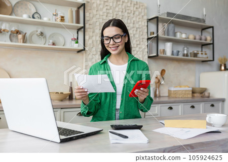 A young woman works and studies at home using a laptop. She is sitting in the kitchen, holding documents in her hands and using the phone. A young woman works and studies at home using a laptop. She is sitting in the kitchen, holding documents in her hands and using the phone. 105924625
