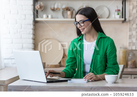 A young woman works and studies at home using a laptop. Sitting in the kitchen at the table and writing in a notebook. 105924633