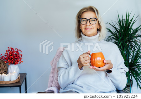 Middle aged woman relaxing with pumpkin shaped cup of hot drink in scandy style hygge interior home with fall mood decor. Lady dreaming, enjoy calm mood without stress, well being alone. Cozy autumn. 105925224