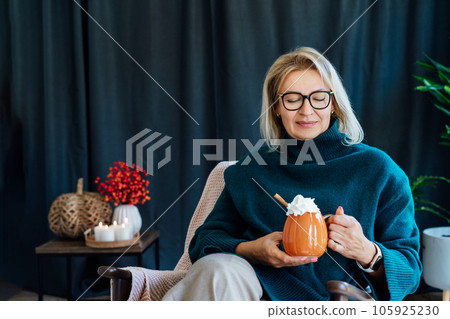 Smiling middle aged woman relaxing with pumpkin shaped cup of hot drink with whipped cream and cinnamon stick with fall mood composition for hygge home decor on background. Cozy autumn holidays 105925230