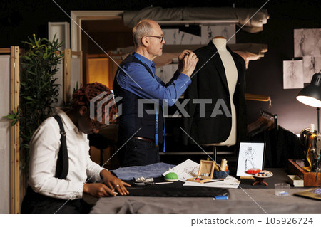 Elderly professional couturier making adjustments on exquisite handmade suit blazer on mannequin while assistant crafts elegant pants. Senior suitmaker and apprentice manufacturing bespoken garments 105926724