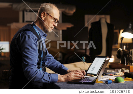 Elderly experienced couturier typing measurements on laptop in professional atelier shop workspace. Skilled expert fashion designer searching online for sartorial inspiration 105926728