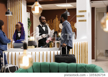 African American hotel receptionist talking with guest, confirming reservation details. Front desk staff checking in tourists arriving at resort during holiday season, customer service in hospitality African American hotel receptionist talking with guest, confirming reservation details. Front desk staff checking in tourists arriving at resort during holiday season, customer service in hospitality 105926734